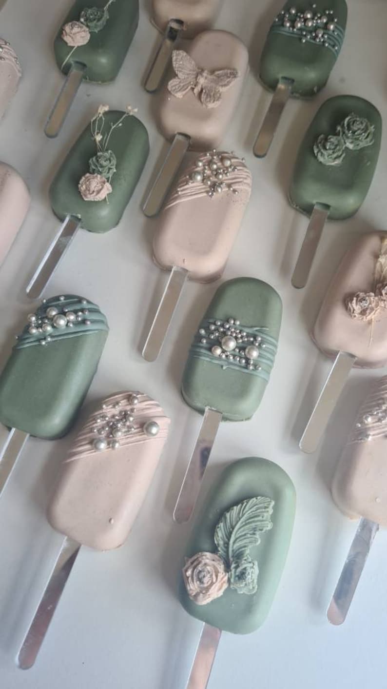 May include: An overhead shot of several pastel-colored cake pops on sticks. Some are sage green, others are light pink. The cake pops are decorated with edible pearls, flowers, and butterfly designs. The pops are arranged on a white surface.