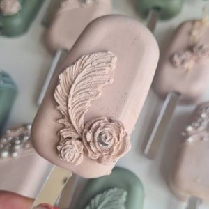 May include: Close-up of several decorative, rectangular treats on sticks. The treats are in shades of pink and green, with floral and feather-like embellishments. One treat is held in a hand with bright pink nail polish.
