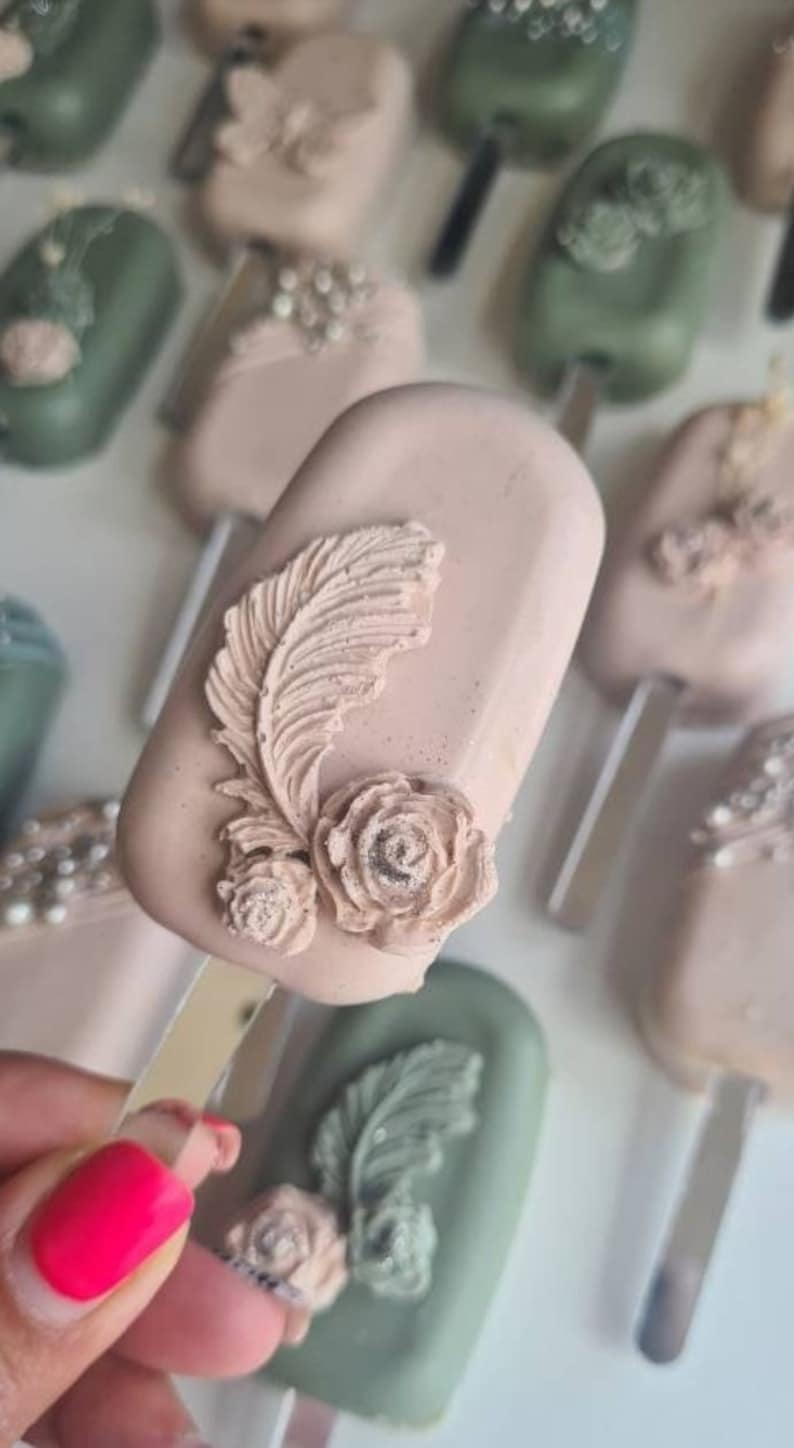 May include: Close-up of several decorative, rectangular treats on sticks. The treats are in shades of pink and green, with floral and feather-like embellishments. One treat is held in a hand with bright pink nail polish.