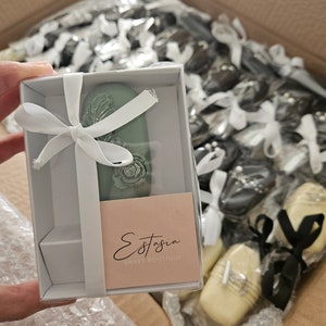 May include: A box of individually wrapped sweets, some in white boxes with white ribbons. One box contains a green oval-shaped sweet with a floral design. The label reads "Estasia Sweet Boutique".