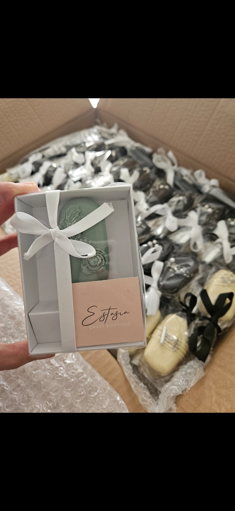 May include: A box of individually wrapped sweets, some in white boxes with white ribbons. One box contains a green oval-shaped sweet with a floral design. The label reads "Estasia Sweet Boutique".