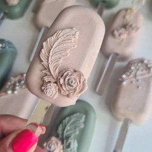 May include: Close-up of several decorative cake pops on sticks. The cake pops are oblong and covered in a light pink and green coating. Each cake pop is decorated with a feather and rose design. Some have pearl accents.