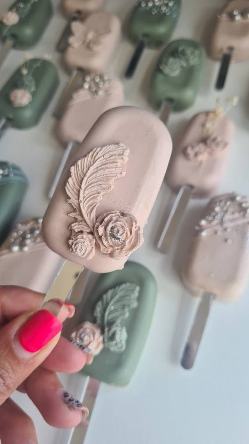 May include: Close-up of several decorative cake pops on sticks. The cake pops are oblong and covered in a light pink and green coating. Each cake pop is decorated with a feather and rose design. Some have pearl accents.