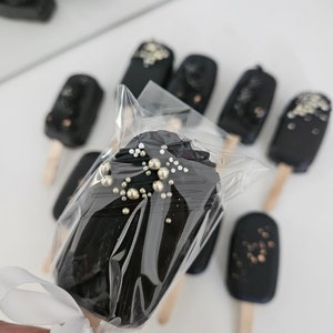 May include: Close-up of several dark chocolate-covered treats on wooden sticks, some with gold and silver sprinkles. One is wrapped in clear plastic and tied with a white ribbon. The treats are arranged on a white surface.