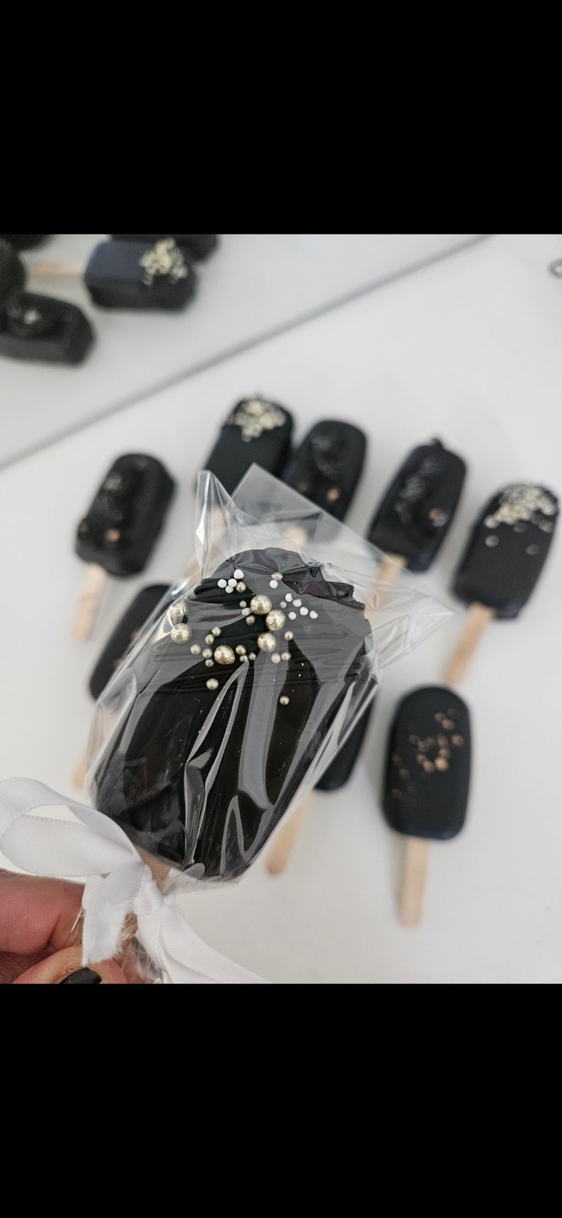 May include: Close-up of several dark chocolate-covered treats on wooden sticks, some with gold and silver sprinkles. One is wrapped in clear plastic and tied with a white ribbon. The treats are arranged on a white surface.