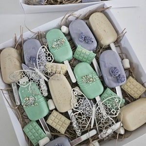 May include: A white box filled with decorated cake pops, chocolate bars, and marshmallows. The cake pops are pastel green, lavender, and beige, adorned with floral and pearl accents. Decorative butterfly cutouts are also included.