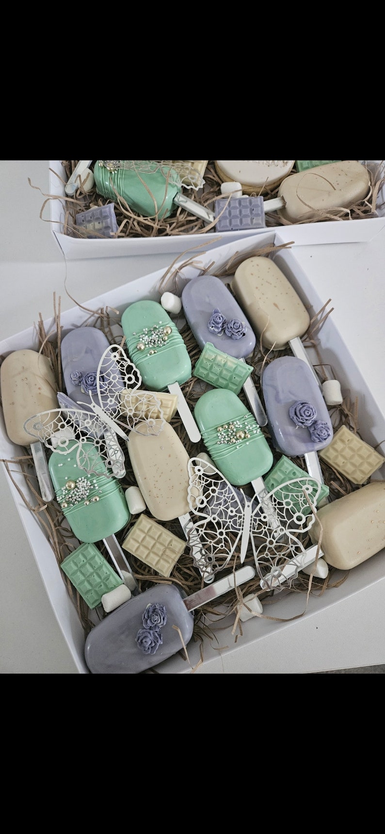 May include: A white box filled with decorated cake pops, chocolate bars, and marshmallows. The cake pops are pastel green, lavender, and beige, adorned with floral and pearl accents. Decorative butterfly cutouts are also included.