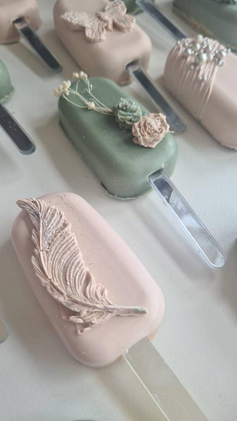 May include: Close-up of several pastel-colored cake pops on sticks. Some are decorated with a butterfly, a feather, and floral accents. The cake pops are arranged on a white surface, creating a visually appealing display.