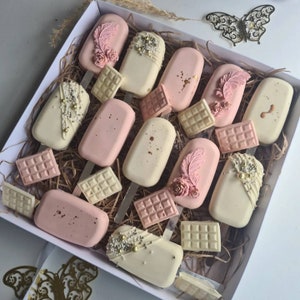 May include: A box filled with decorated ice cream bars and chocolate bars. The ice cream bars are white and pink, adorned with edible decorations like pearls, feathers, and flowers. The chocolate bars are in shades of white and pink.