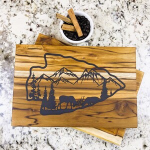 Peut inclure: Une planche à découper rectangulaire en bois avec une incrustation sombre en forme de pointe de flèche. L'incrustation représente une chaîne de montagnes, des conifères et deux élans broutant dans une forêt. Le grain du bois est visible sur toute la planche.