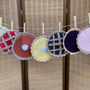 May include: A set of seven circular crochet coasters hanging from a white string with wooden clothespins. The coasters feature various designs including solid colors in light green, rust, dark purple, and light pink, alongside pie-themed patterns.