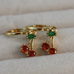 May include: A pair of gold hoop earrings with a dangling cherry charm. The cherry is made of red and green cubic zirconia stones.