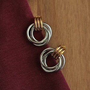 Peut inclure: Une paire de boucles d'oreilles argentées avec un accent doré. Chaque boucle d'oreille comporte trois anneaux entrelacés et une tige dorée. Les boucles d'oreilles sont présentées sur un tissu bordeaux et une surface en bois.