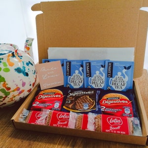 May include: A brown cardboard gift box filled with a variety of snacks, including tea bags, biscuits, and chocolate. The box is open and the snacks are visible. The box is sitting on a wooden surface. The text on the tea bags reads "A bag of our organic decaf tea". The text on the biscuits reads "Digestives 2 Wheatmeal Biscuits" and "Lotus Biscoff Happy to see you again".