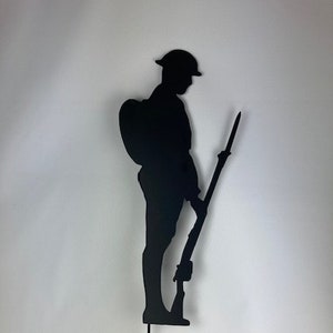 May include: Black metal silhouette of a soldier holding a rifle, standing to attention. The silhouette is mounted on a metal stake.