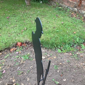 May include: Black metal silhouette of a person standing in a grassy area. The silhouette is of a person in a military uniform, possibly a soldier.