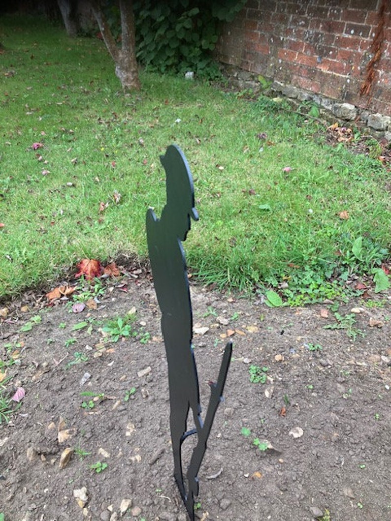 May include: Black metal silhouette of a person standing in a grassy area. The silhouette is of a person in a military uniform, possibly a soldier.