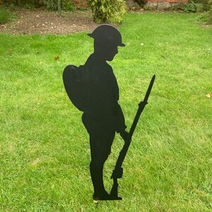 May include: A black metal silhouette of a soldier standing in a grassy field. The soldier is wearing a helmet and carrying a rifle.
