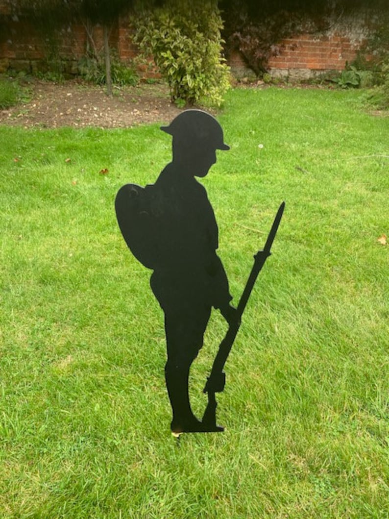 May include: A black metal silhouette of a soldier standing in a grassy field. The soldier is wearing a helmet and carrying a rifle.