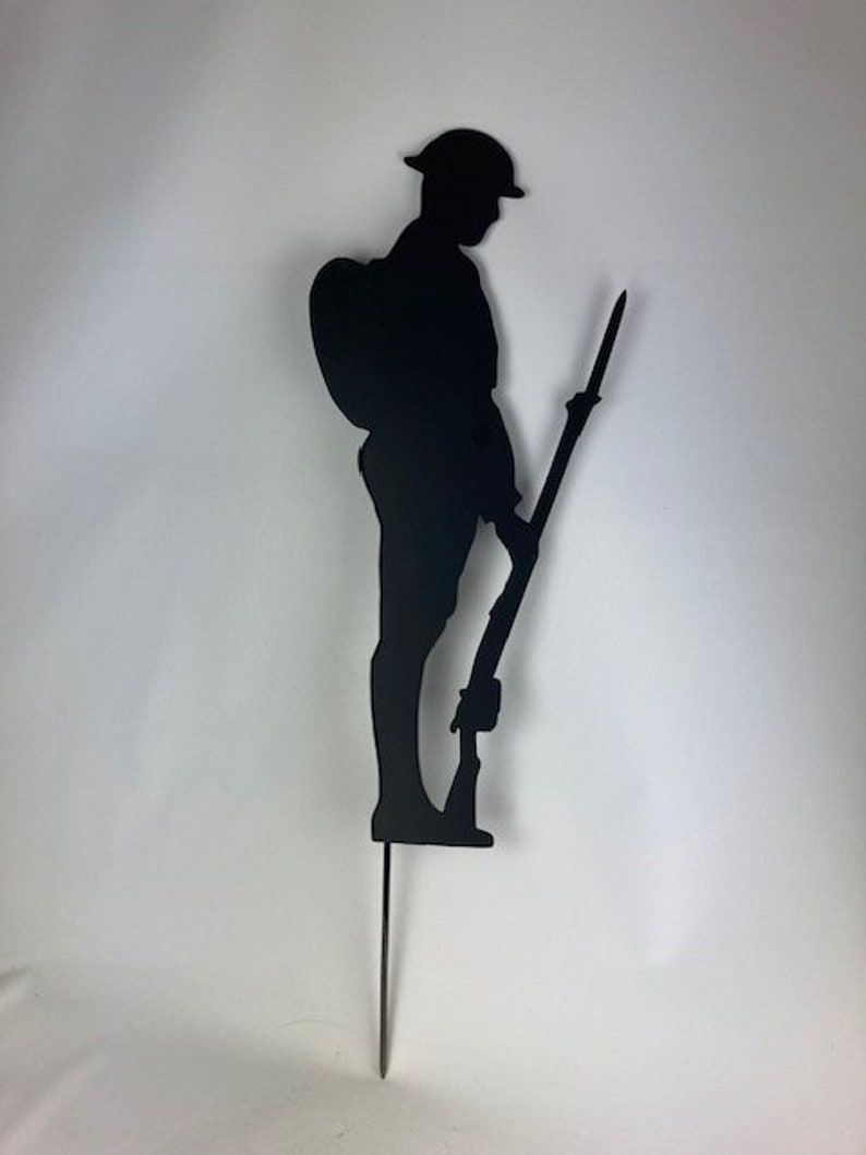 May include: Black metal silhouette of a soldier holding a rifle, standing to attention. The silhouette is mounted on a metal stake.
