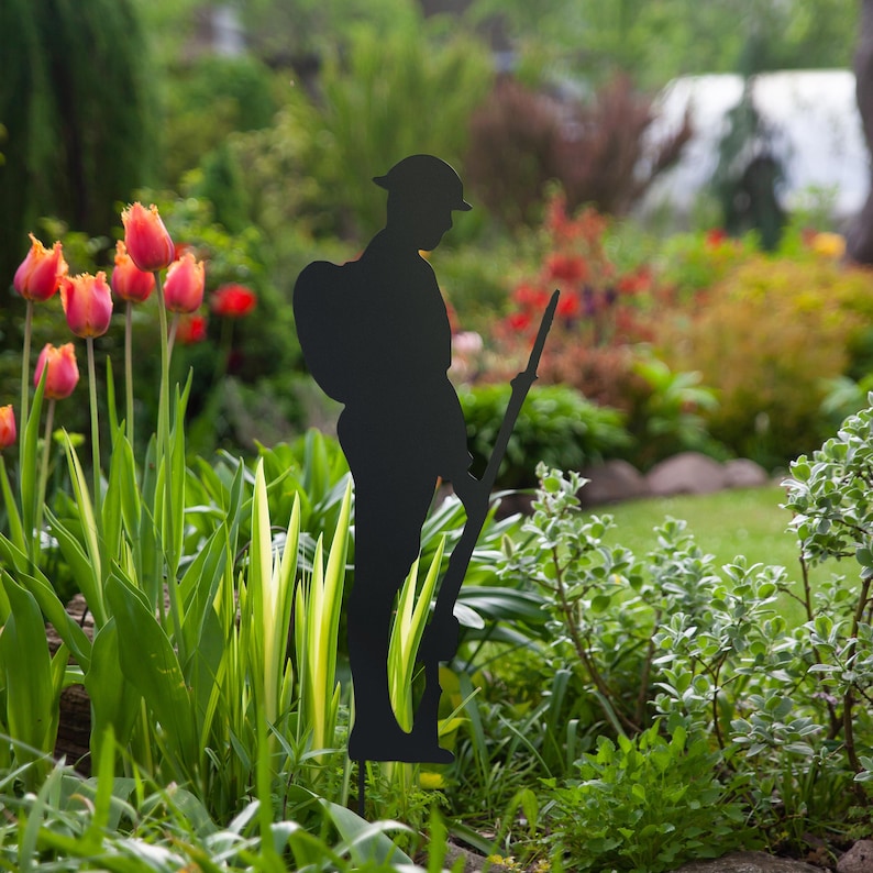 May include: Black metal silhouette of a soldier standing to attention, holding a rifle. The silhouette is placed in a garden setting with green grass and red tulips.