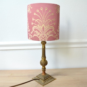 Pode incluir: Uma lâmpada de mesa vintage com uma cúpula rosa com um padrão floral creme. A lâmpada tem uma base e haste cor de latão e uma base quadrada. A lâmpada tem aproximadamente 30 cm de altura.