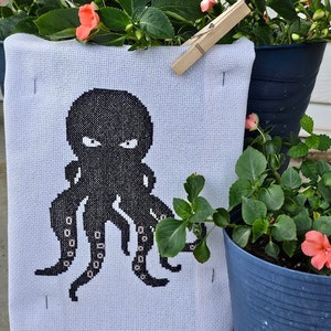 以下が含まれることがあります： 黒いタコが8本の触手で描かれた白いクロスステッチ刺繍。刺繍は木製の洗濯ばさみで壁に掛けられている。刺繍はピンクの花が咲いた緑の植物に囲まれている。