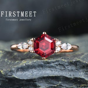 May include: A rose gold ring featuring a hexagonal red gemstone surrounded by smaller white gemstones. The ring is displayed on a grey textured surface.
