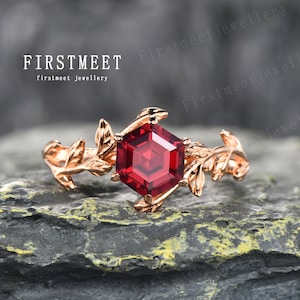 May include: A rose gold ring featuring a hexagonal red gemstone, surrounded by delicate leaf details. The ring is displayed on a dark grey textured surface. FIRSTMEET firstmeet jewellery.