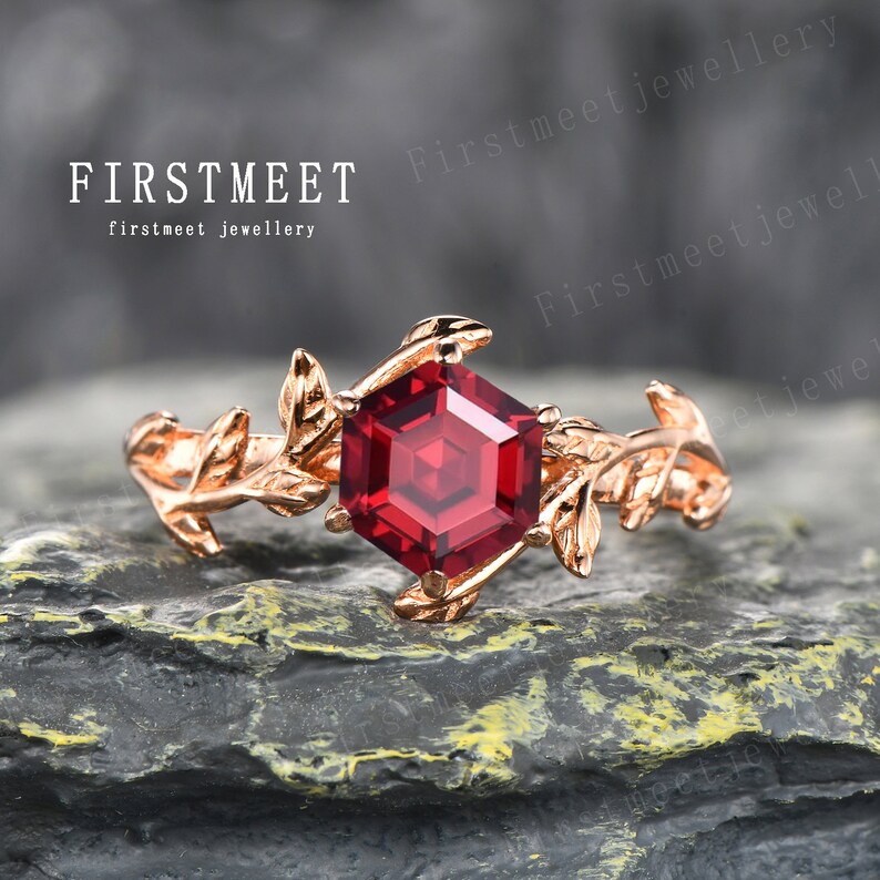 May include: A rose gold ring featuring a hexagonal red gemstone, surrounded by delicate leaf details. The ring is displayed on a dark grey textured surface. FIRSTMEET firstmeet jewellery.