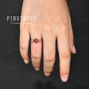 May include: A gold ring with a red gemstone set in a square shape. The ring is on a finger and the background is black.