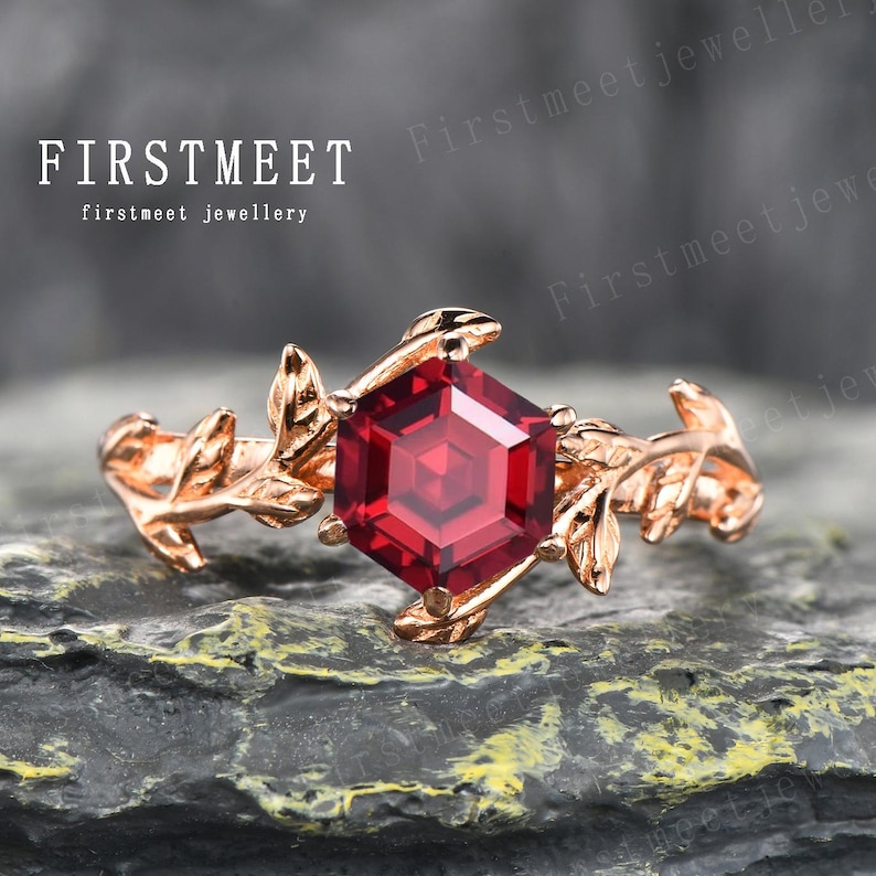 May include: A rose gold ring featuring a hexagonal red gemstone, surrounded by delicate leaf details. The ring is displayed on a textured grey surface.
