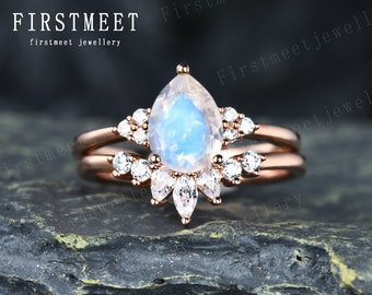 May include: A rose gold engagement ring set with a pear-shaped moonstone centre stone and a halo of smaller round stones. The ring is set on a textured grey surface.