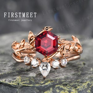 Op de afbeelding: Een verlovingsring van roségoud met een rode edelsteen in een zeshoekige vorm. De ring is versierd met een delicaat bladontwerp en kleinere witte edelstenen.