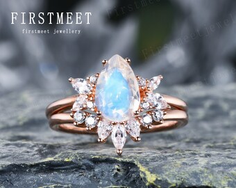 May include: A pear-shaped moonstone engagement ring set in rose gold with a halo of smaller diamonds. The ring is set on a grey rock.
