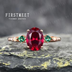 Puede incluir: Un anillo de oro rosa con un gran rubí ovalado como piedra central, flanqueado por esmeraldas cuadradas y pequeños acentos de ópalo. El anillo se muestra sobre un fondo oscuro y texturizado. El texto "FIRSTMEET firstmeet jewellery" es visible.