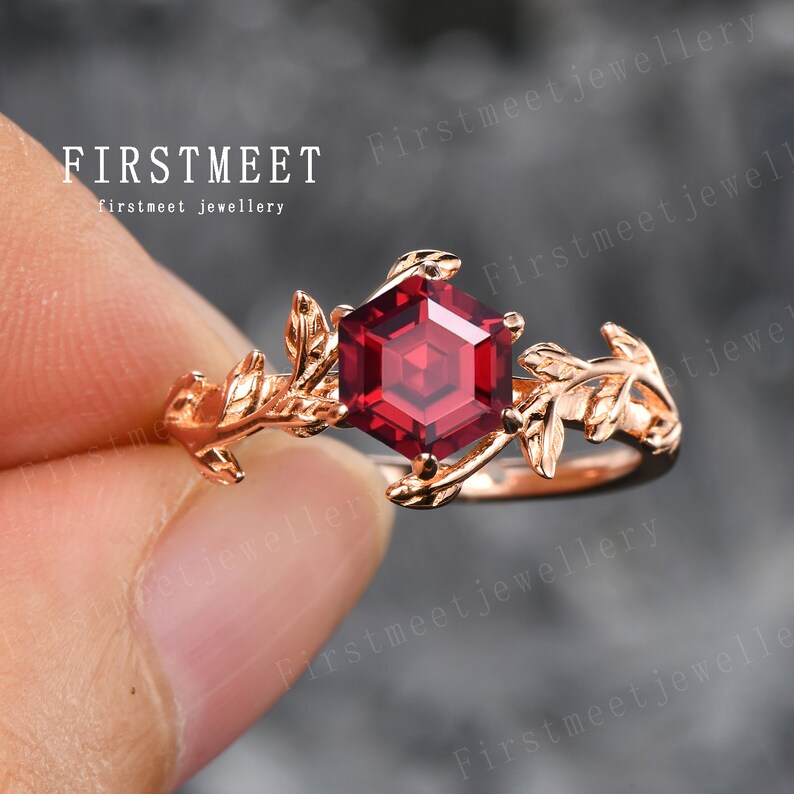 May include: A rose gold ring featuring a red gemstone and leaf details. The ring is displayed on a finger.