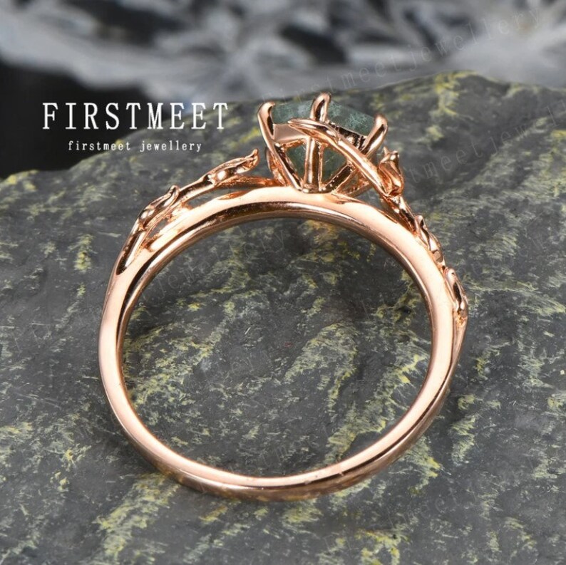 May include: A rose gold ring with a green gemstone set in a delicate floral design. The ring is on a textured grey surface. FIRSTMEET jewellery.