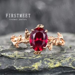 Puede incluir: Un anillo de oro rosa con una gran piedra de rubí ovalada rodeada de delicados detalles de hojas. El anillo está colocado sobre una superficie gris texturizada.