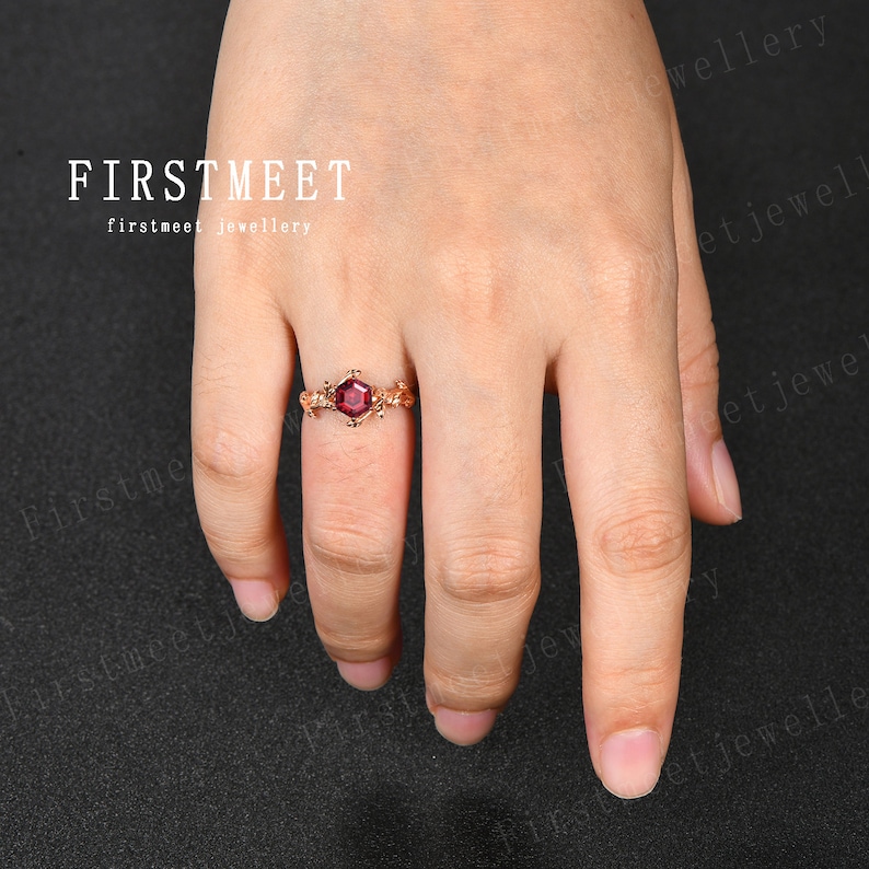 May include: A gold ring with a red gemstone set in a square shape. The ring is on a finger and the background is black.