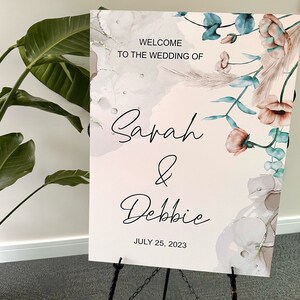 May include: Welcome sign for a wedding with a floral design and the names Sarah & Debbie. The sign is on a black easel and the date July 25, 2023 is printed below the names.