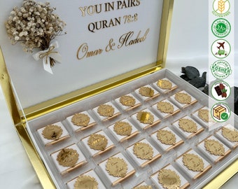 May include: A white and gold gift box with chocolates. The box lid has gold lettering that reads "AND WE CREATED YOU IN PAIRS" and "QURAN 78:8". The chocolates are individually wrapped with gold labels that read "Omar & Hadeel".