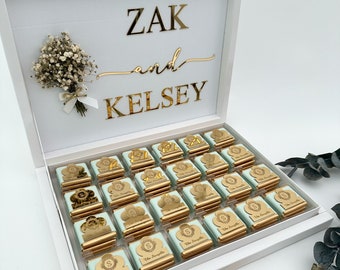 以下が含まれることがあります： 白いギフトボックスに「ZAK and KELSEY」という金色の文字が書かれています。ボックスには、薄い緑色と金色のホイルで包まれた小さな正方形のチョコレートが入っています。チョコレートにはそれぞれ金色の「S」が書かれています。