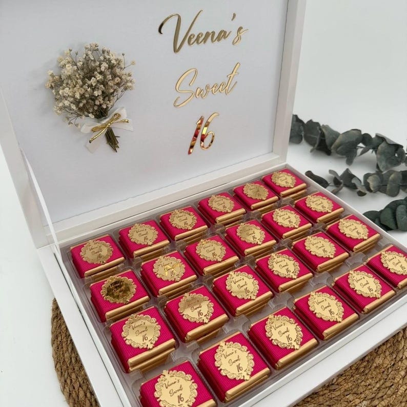 May include: A white box with a clear lid, filled with pink wrapped chocolates. The box is decorated with gold text that reads "Veena's Sweet 16". A small bouquet of dried flowers and a gold bow are attached to the box.