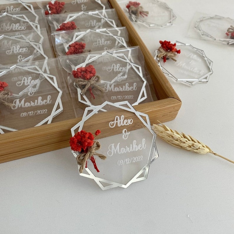 K&ouml;nnte beinhalten: Ein Set aus klaren Acryl-Hochzeitsgeschenken mit geometrischem Design. Jedes Geschenk tr&auml;gt die Namen "Alex & Maribel" und das Datum "09/12/2022" in wei&szlig;er Gravur. Die Geschenke sind mit einem kleinen Strau&szlig; getrockneter roter Blumen verziert.