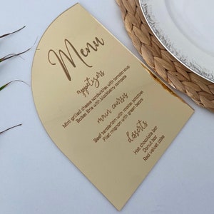 May include: A gold-colored menu card with a rounded top. The menu is written in a cursive font and includes appetizers, main courses, and desserts. The menu items include mini grilled cheese sandwiches, baked brie, beef tenderloin, filet mignon, hot chocolate bar, donut bar, and red velvet cake.