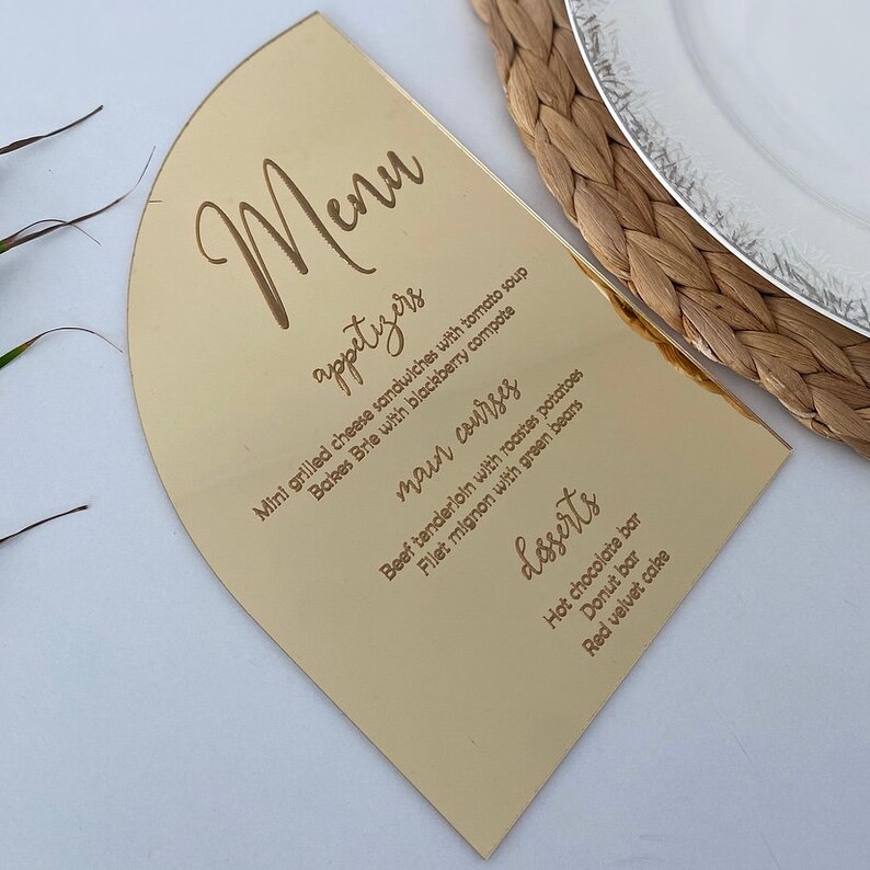 May include: A gold-colored menu card with a rounded top. The menu is written in a cursive font and includes appetizers, main courses, and desserts. The menu items include mini grilled cheese sandwiches, baked brie, beef tenderloin, filet mignon, hot chocolate bar, donut bar, and red velvet cake.