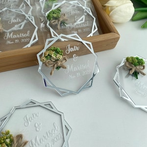K&ouml;nnte beinhalten: Silberne Spiegel-Acryl-Hochzeitsgeschenke mit geometrischem Design. Jedes Geschenk enth&auml;lt einen kleinen Strau&szlig; getrockneter Blumen und die Namen des Paares, Jair & Marisol, sowie das Hochzeitsdatum, den 19. November 2022.