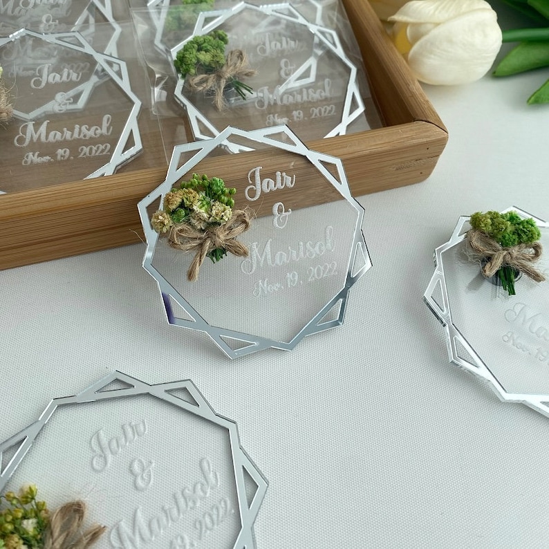 K&ouml;nnte beinhalten: Silberne Spiegel-Acryl-Hochzeitsgeschenke mit geometrischem Design. Jedes Geschenk enth&auml;lt einen kleinen Strau&szlig; getrockneter Blumen und die Namen des Paares, Jair & Marisol, sowie das Hochzeitsdatum, den 19. November 2022.