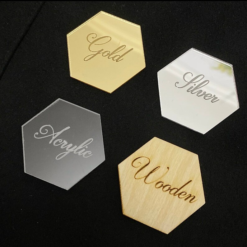 May include: Four hexagonal place cards made of different materials: gold, silver, acrylic, and wood. Each card has the material name written in cursive script.